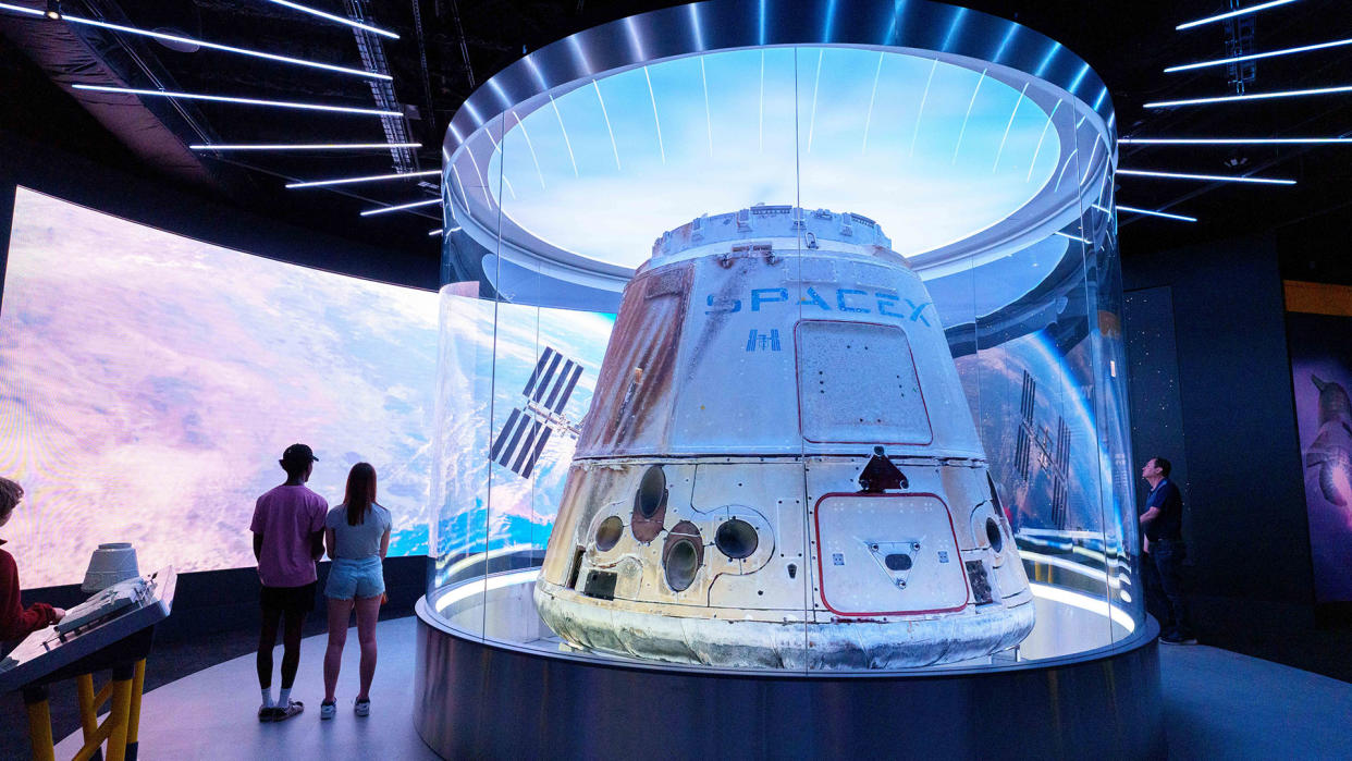  A white space capsule on display in a museum, with several people nearby. 