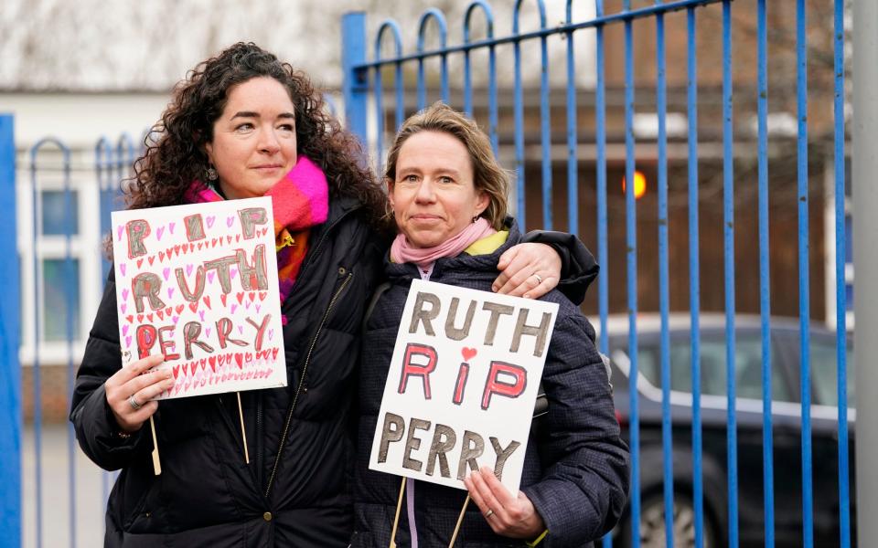 Two women stand outside a school holding home-made placards saying 'RIP Ruth Perry'