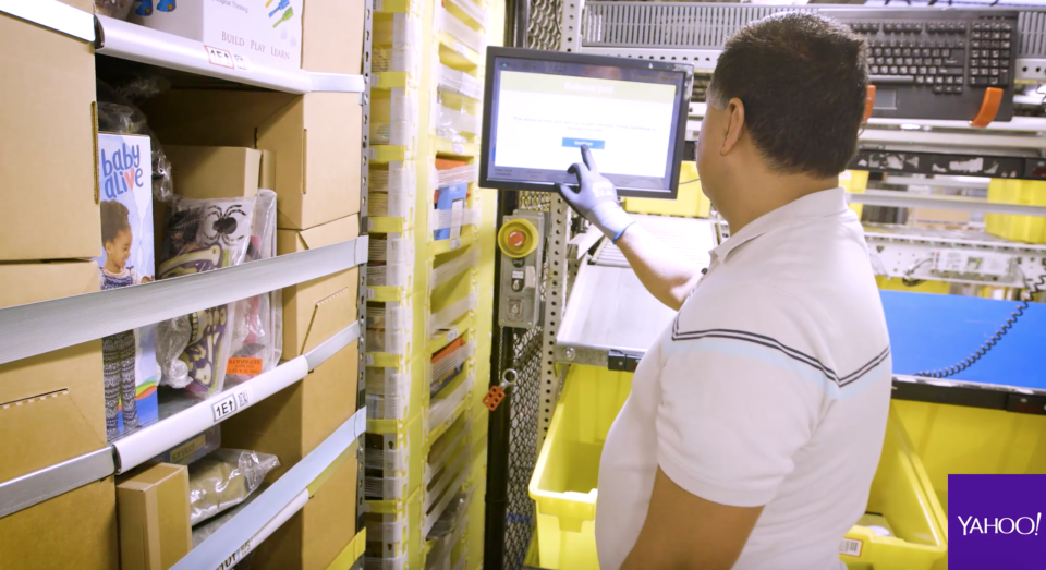 An Amazon employee waiting at a “pick station” to pick an item from a shelf and put it in a yellow bin, or “tote.” Source: Yahoo Finance