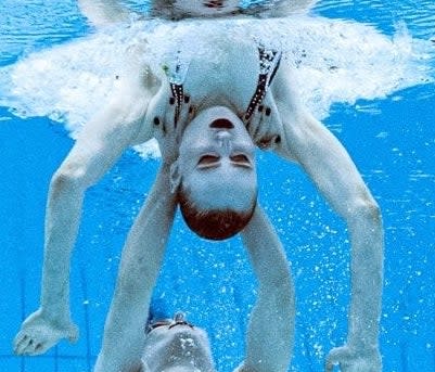 A swimmer underwater appearing to hold up another one, who's upside down, at the shoulders