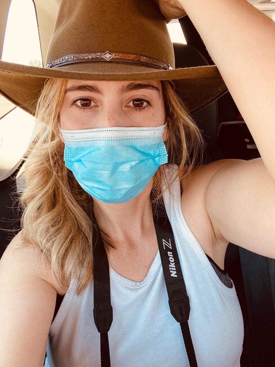 A woman wearing a cowboy hat and a mask takes a selfie inside a car.