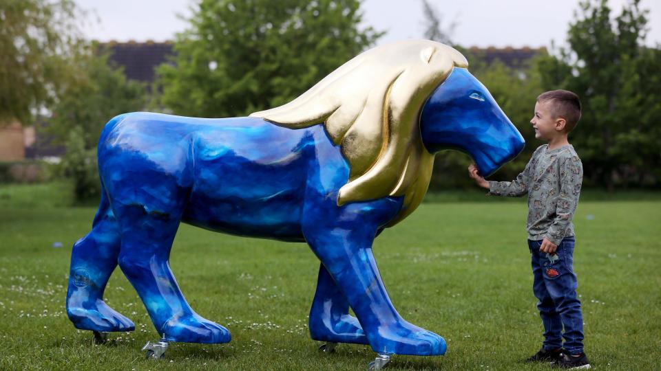 A boy smiles as he touches the chin of a blue lion sculpture with a gold mane in a park