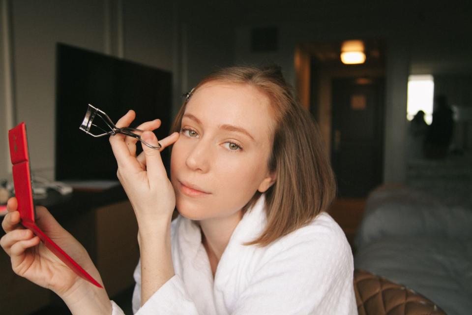 hannah einbinder holding an eyelash curler and compact and gazing at the camera