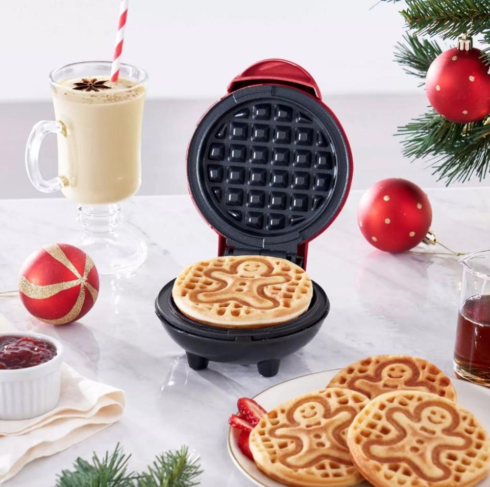 Waffle maker open with gingerbread-shaped waffle inside, next to plate of waffles, mug of eggnog, and red ornaments on white tablecloth