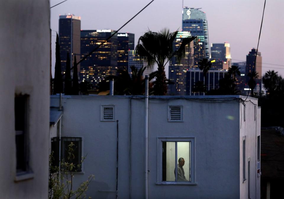 A man is framed in the window of an apartment, with a downtown L.A. in the distance.