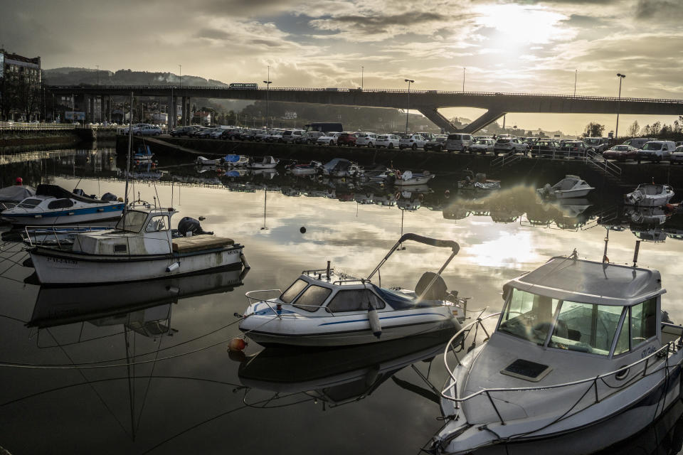 Small pleasure boats are moored in a row on the Ria de Pontevedra, with an elevated freeway and parking lot in the distance.