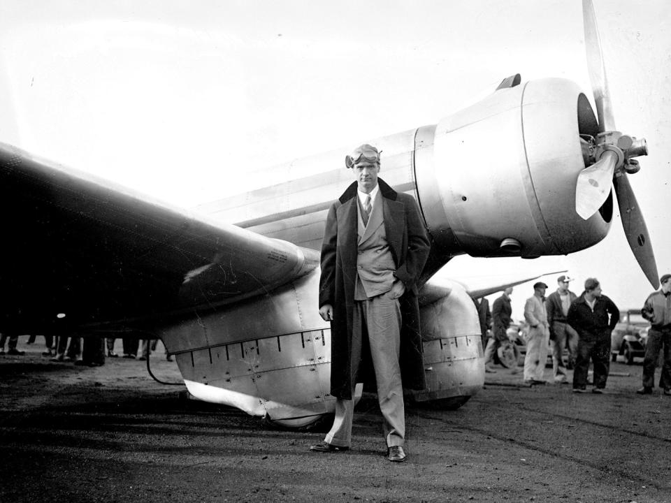 Howard Hughes standing in front of a plane wearing aviator goggles.