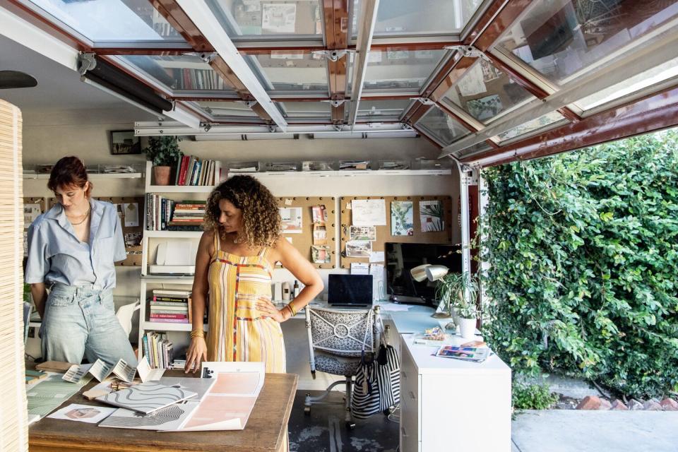 Two women stand in a garage workspace, looking at papers on a desk.