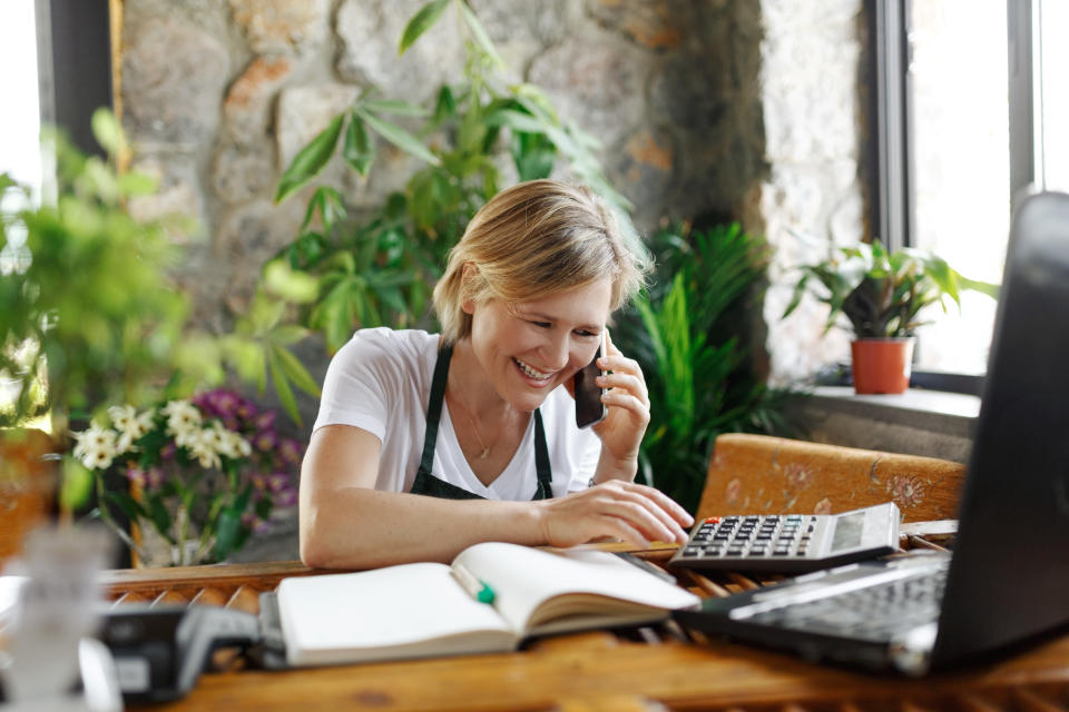 Young woman entrepreneur sitting at desk and typing on computer while chatting on smartphone. Happy female florist in green floral center tapping on laptop and counts on calculator.