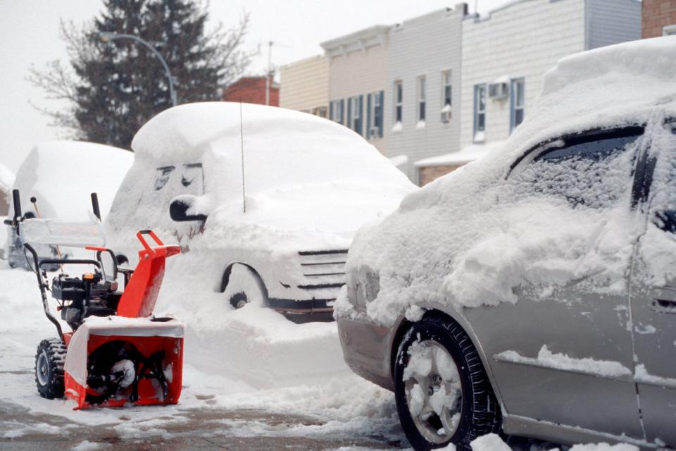 Red snow blower parked on sidewalk next to snow-covered cars. 