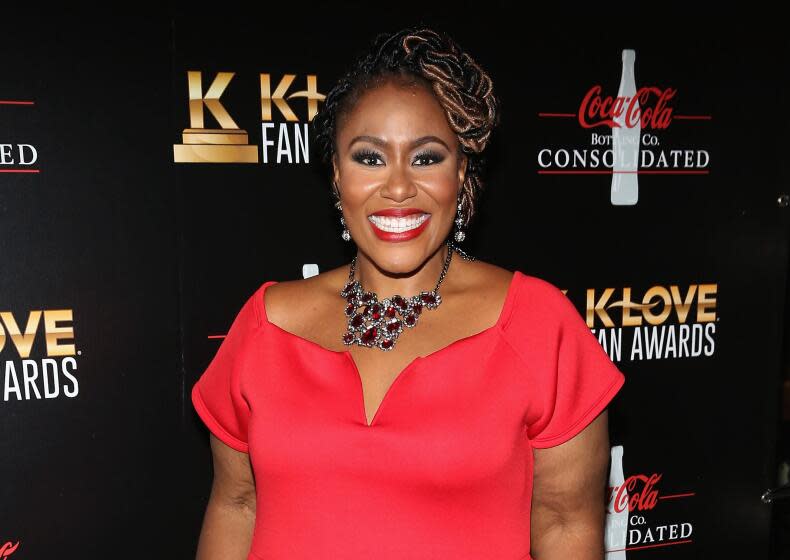 A Black woman in a red gown with hair in a braided style and a necklace smiling against a black background