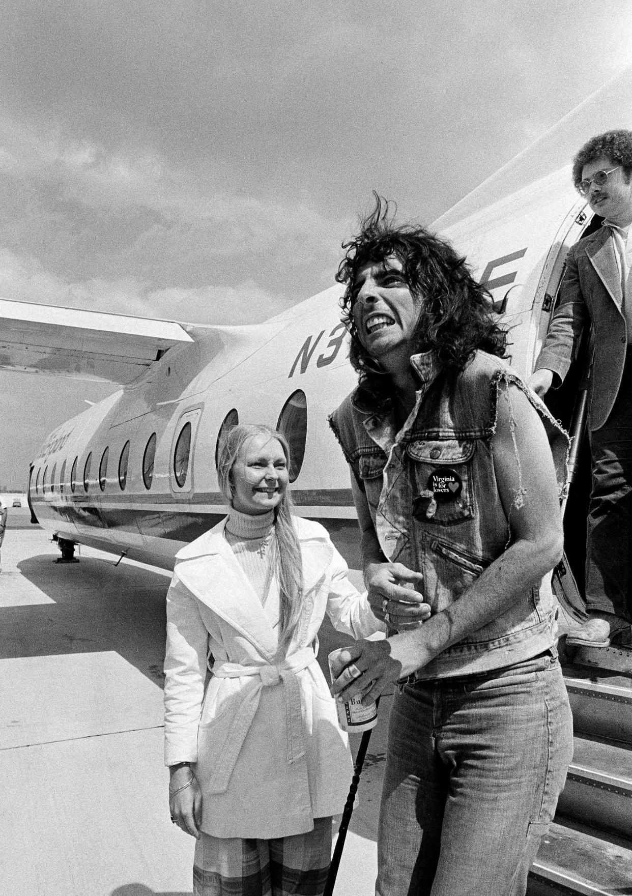 Glam rock star, Alice Cooper, right, reacts to the cold weather as he arrives at Logan International Airport in Boston, Mass., on a chartered Air New England flight from Philadelphia, April 26, 1975. Hostess Roberta Anderson sees Cooper off the plane. Cooper and his band will perform at Boston Garden. (AP Photo/J. Walter Green)