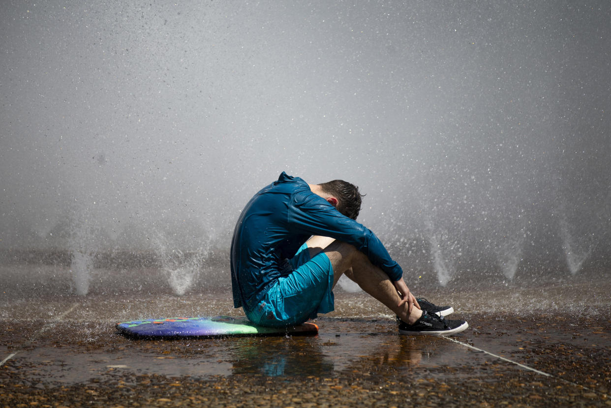 Sitting on the wet ground surrounded by sprinklers, a man dressed in shorts and a long shirt soaks in the heat.