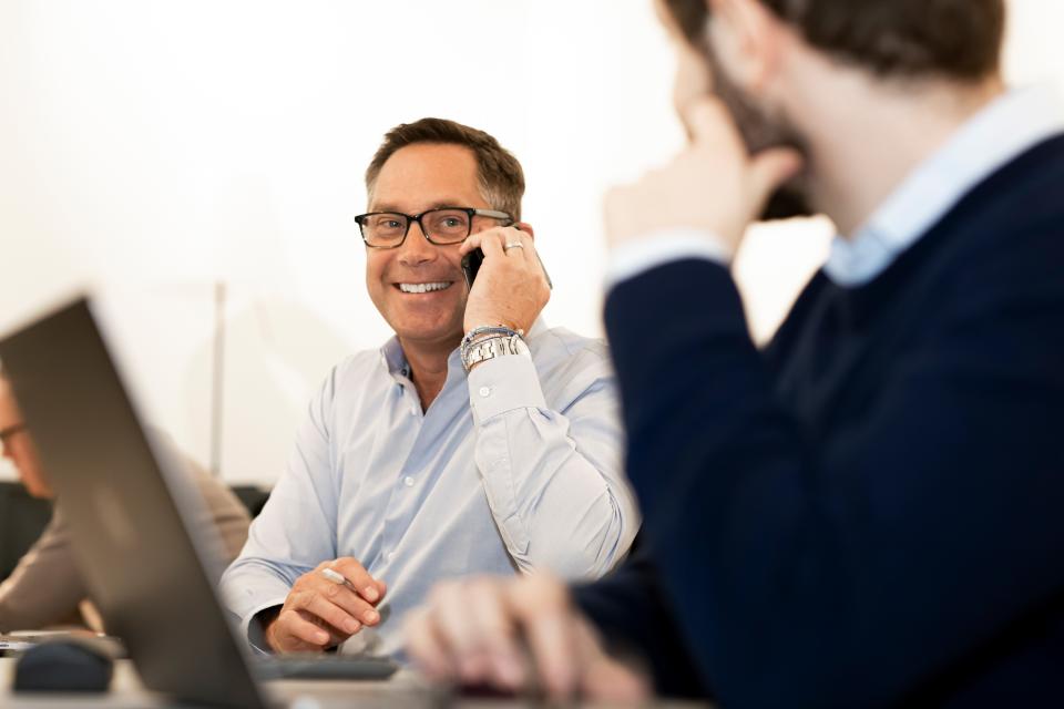 mark bula wearing a button down and black glasses holds his phone up to his ear as he smiles at a colleague who's back is facing the camera. behind bula is H2 Green Steel's CEO Henrik Henriksson