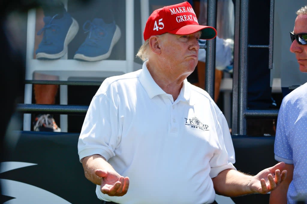 Trump, wearing a white shirt and a MAGA hat, holds his hands with his palms up while in conversation with a man to his left.