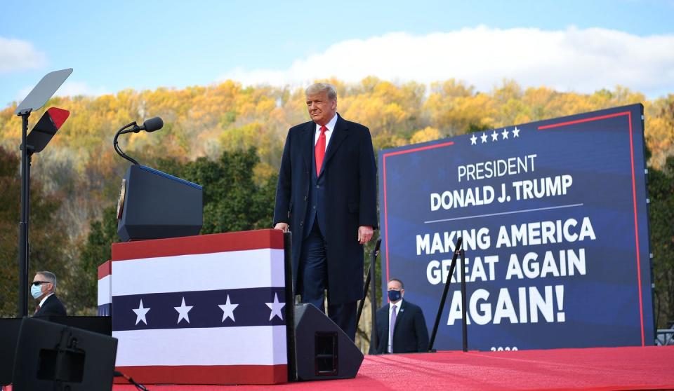 A man in a suit and overcoat stands in front of a sign saying 