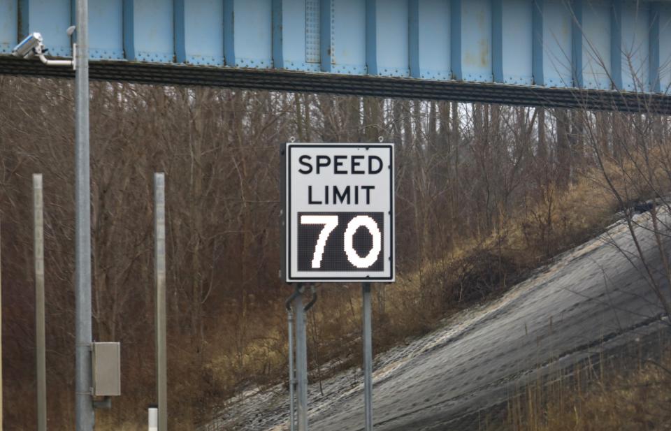 Speed limit sign displaying 70 below a road bridge with a hillside and trees in the background