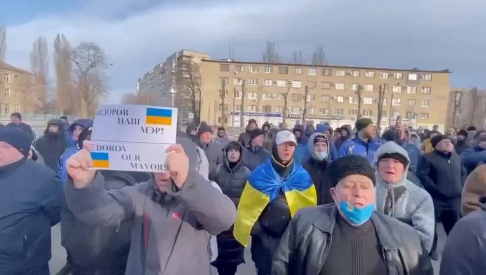 A crowd of Ukrainian men stand outside in Melitopol holding up signs in protest of the abduction of their mayor.