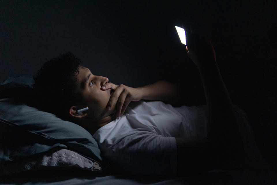 A person lies in bed in a dark room with a hand on their chin and looks at a smartphone, wearing a white shirt and white wireless earbuds