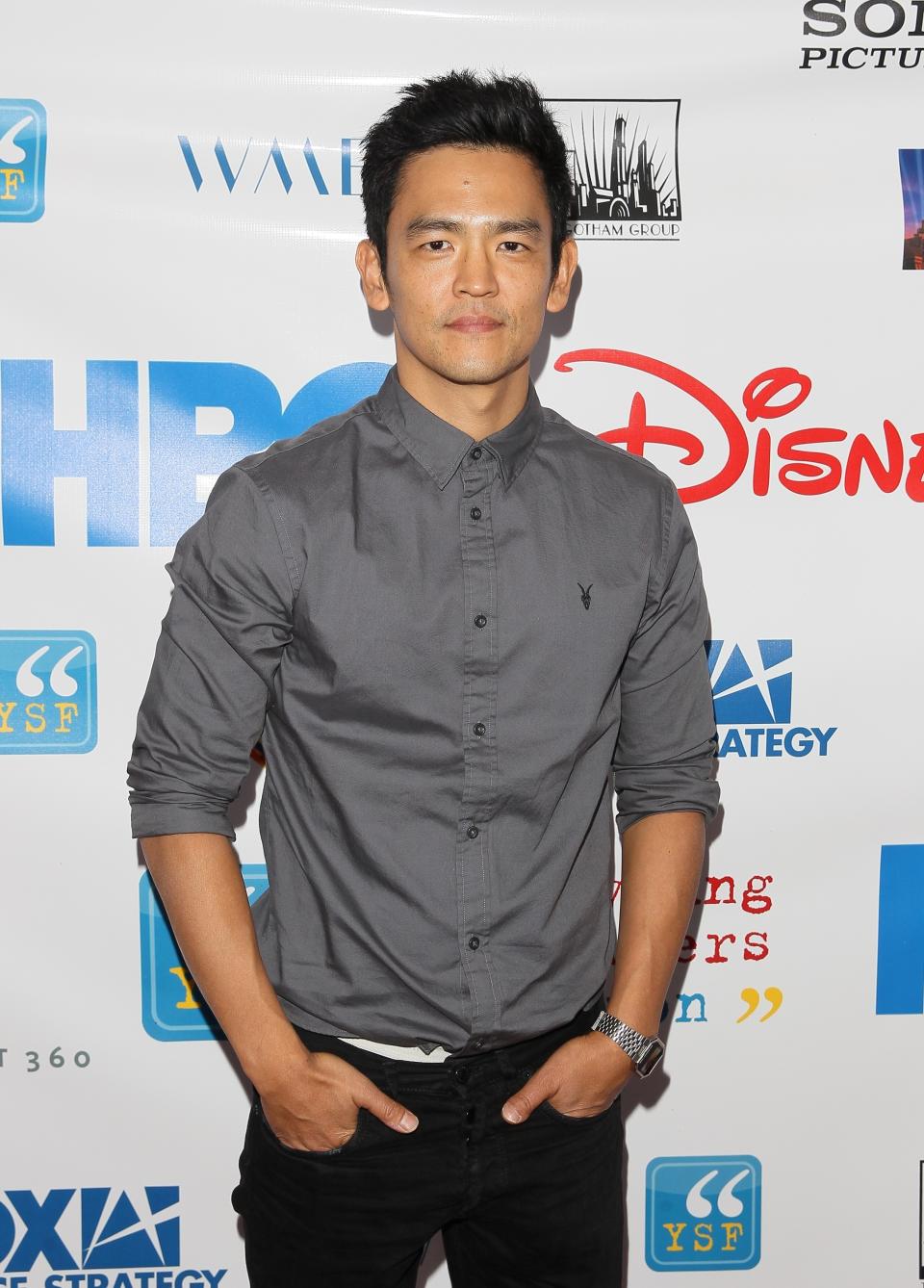 John Cho in a casual button-up shirt and dark jeans, posing on the red carpet in front of HBO, Disney, and Sony logos