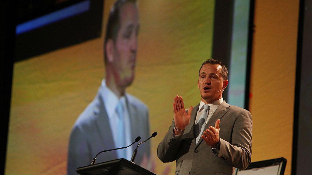 A man in a light coloured suit stands in front of a lectern delivering a speech into a microphone. He also appears enlarged on the big screen behind him.