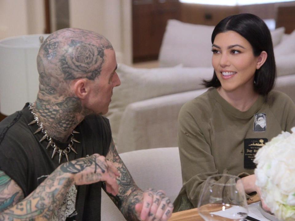 travis barker and kourtney kardashian sit together at a table, smiling at each other. there are flowers visible on the table in front of them