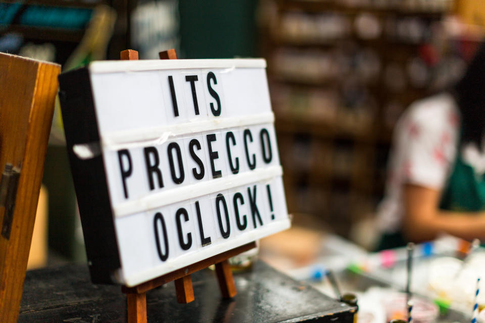 Close up color image of a fun handmade wooden sign with the words 'It's Prosecco o'clock'. The image was taken at a food and drink market in London, UK. In the background the market, and a female figure working on the stall, is defocused, leaving room for copy space.