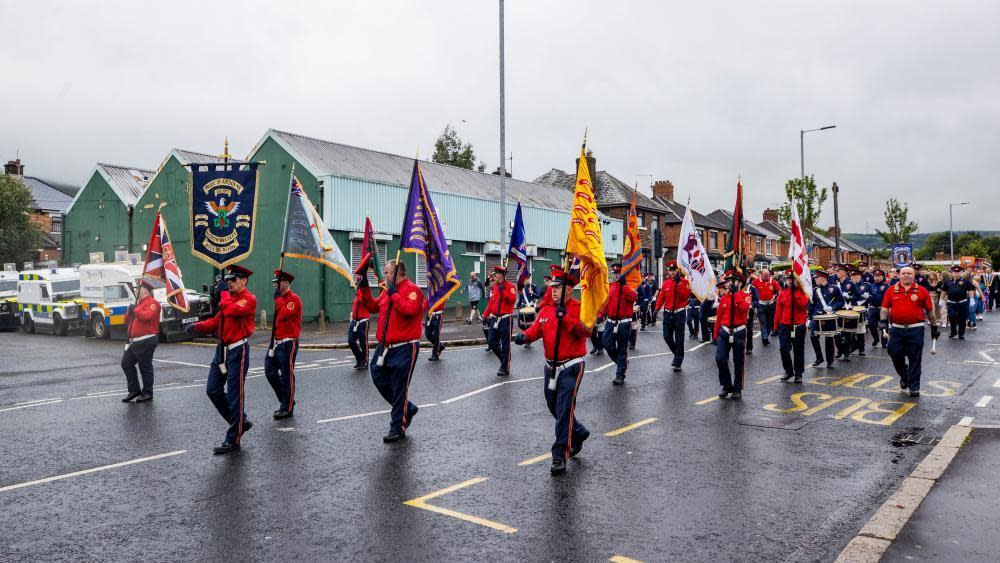 Members of a flute band wearing red uniforms, holding flags, walk along a road with police land rovers in the background. 