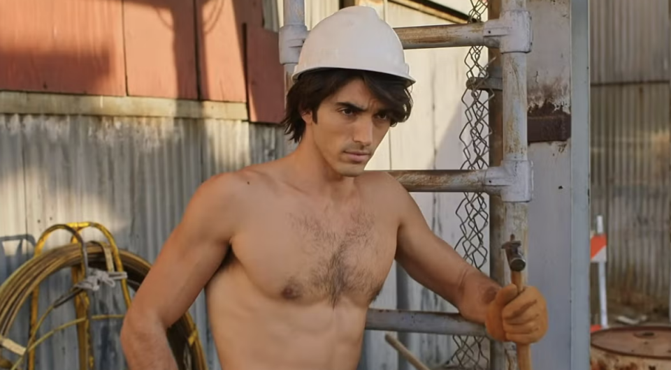 A person wearing a white construction helmet and gloves stands shirtless at a work site with metal structures and coiled cables visible in the background