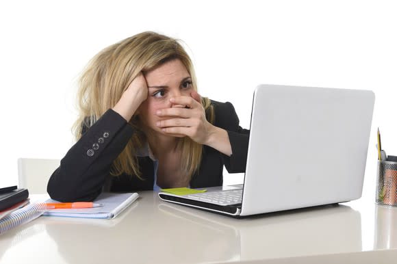 A woman looking at a laptop with a disturbed expression while covering her mouth with her left hand.