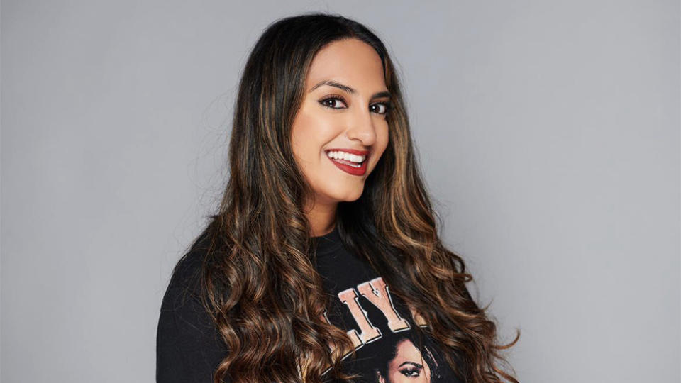 Jasmine Takhar, an Asian woman, smiling. She is wearing a black jumper with pink writing partially visible and partly covered by her hair. The background is plain grey.