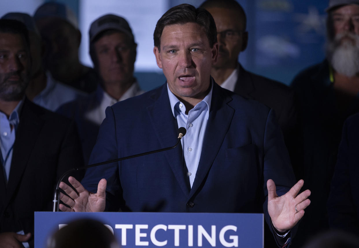 Florida Gov. Ron DeSantis at the podium, above a sign that begins: Protecting...