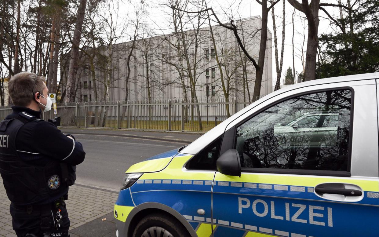 A policeman in black stands with arms folded and back to the camera facing a brutal grey cube of a building behind a spiky metal fence