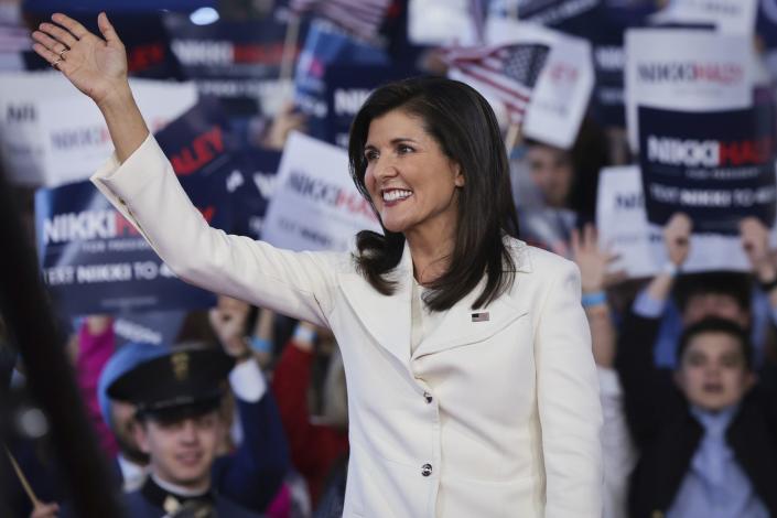 nikki haley wearing a red jacket and smiling while waving at supporters at a campaign rally, with spectators holding up signs and cheering in the background