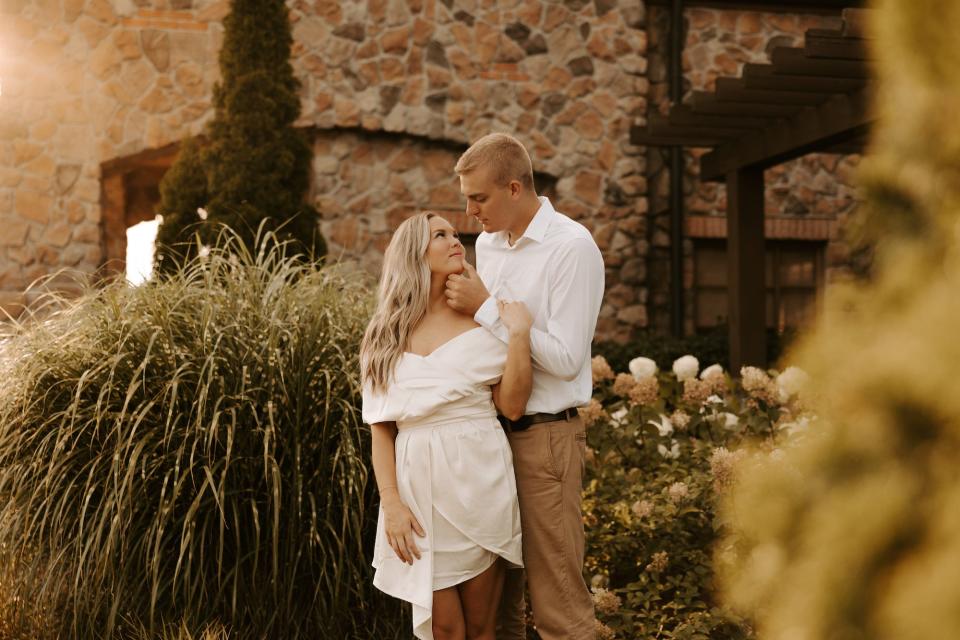 A woman in a white dress and a man in a white shirt and khakis look at each other as he holds her chin in front of a stone building.