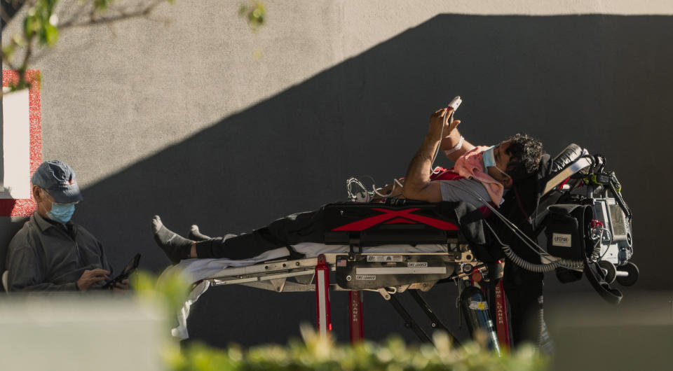 An unidentified patient uses his mobile phone while receiving oxygen on a stretcher, as Los Angeles Fire Department Paramedics monitor him outside the Emergency entrance, waiting for his room at the CHA Hollywood Presbyterian Medical Center in Los Angeles Friday, Dec. 18, 2020. Increasingly desperate California hospitals are being "crushed" by soaring coronavirus infections, with one Los Angeles emergency doctor predicting that rationing of care is imminent. (AP Photo/Damian Dovarganes)