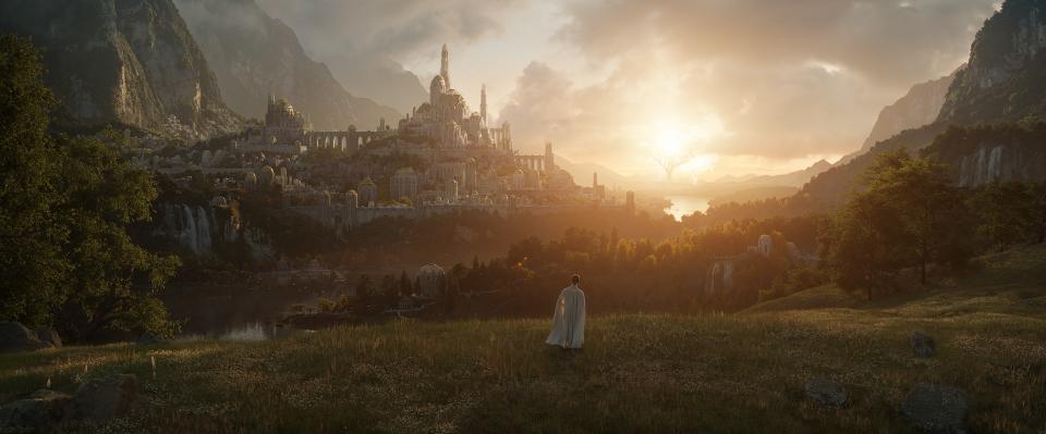 A figure wearing white stands, very small in the image, on a green pasture, facing a massive white kingdom of Middle-earth in The Lord of the Rings Amazon series.