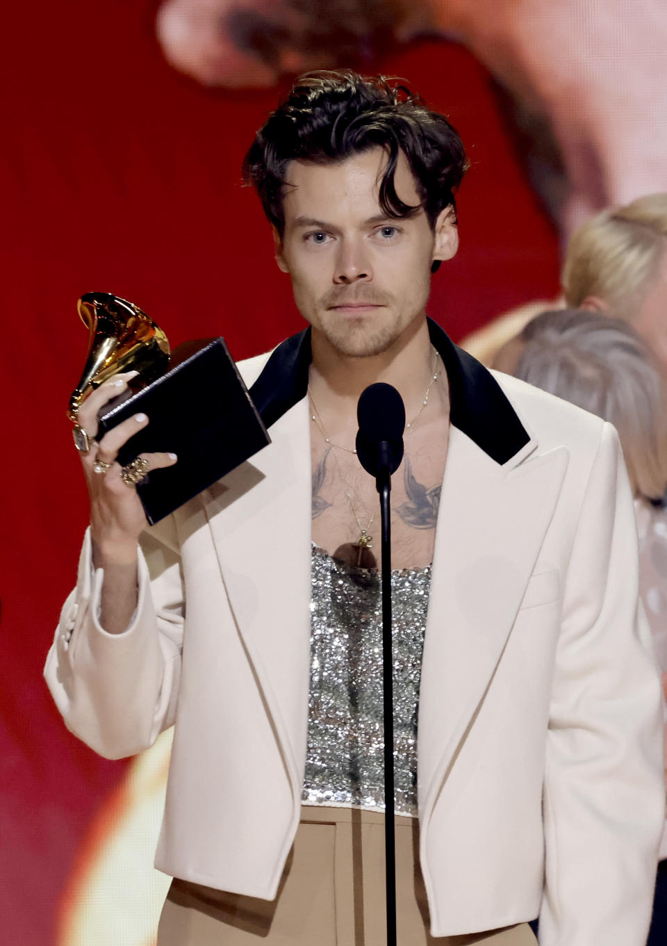 Harry Styles in a cream suit jacket with a glittering shirt, holding a Grammy award onstage