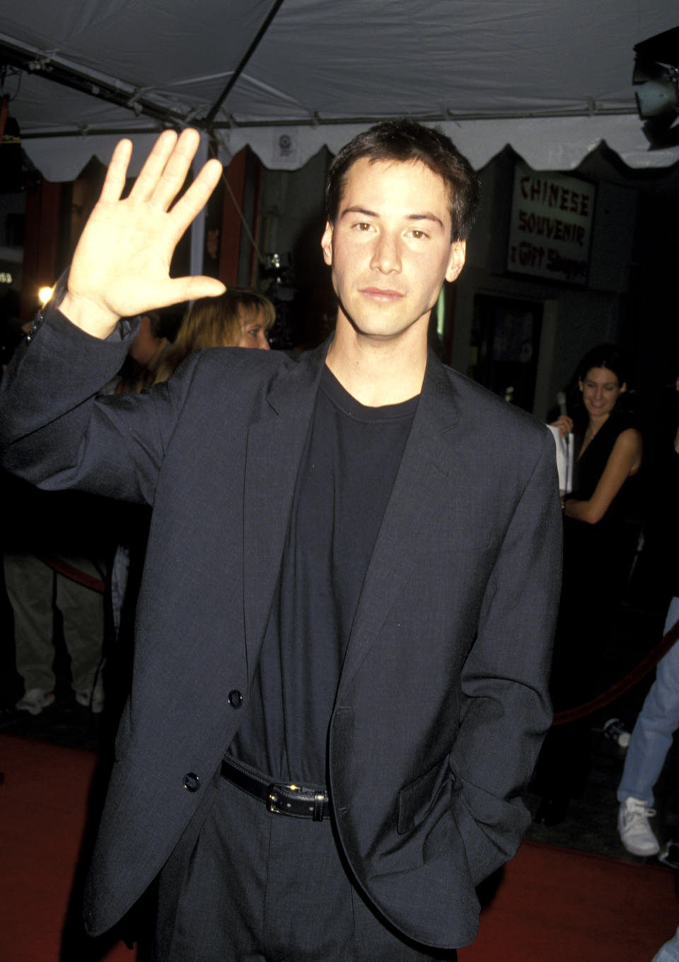 Keanu Reeves waves on the red carpet, wearing a casual dark suit