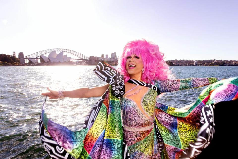 A smiling drag queen in a pink wig stands in front of the Sydney Opera House