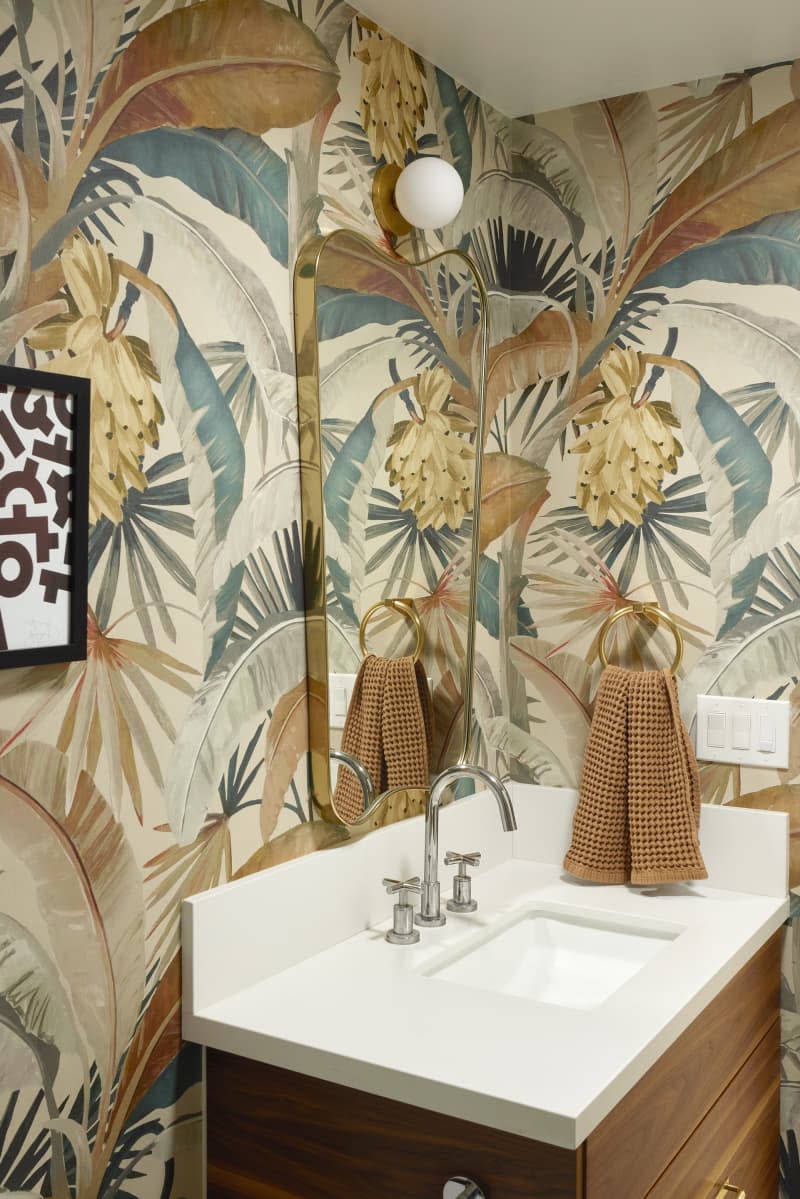 beige and blue tropical floral wallpaper, vintage mirror and lighting fixture, white countertop with wood vanity underneath.