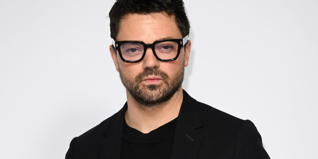 dominic cooper, a man stands looking at the camera with a stern facial expression, he has short black hair and a beard, he wears thick black glasses, a black tshirt with black trousers and jacket