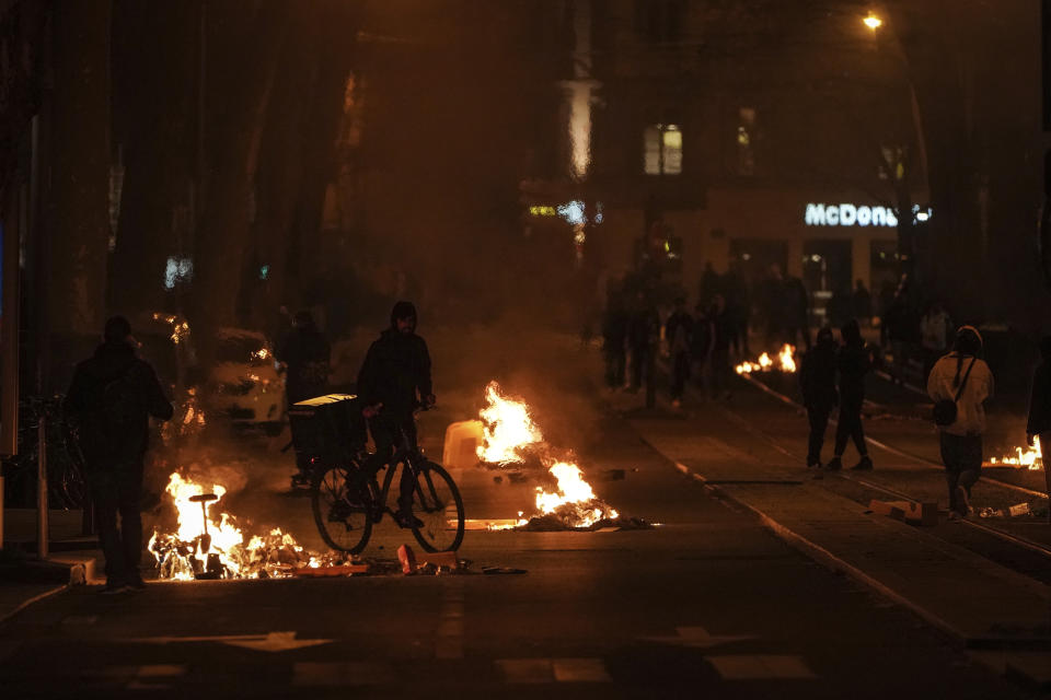 A man passes by fires set by protesters in Lyon, central France, after parliament adopted a divisive pension bill, Monday, March 20, 2023. The French government has survived two no-confidence votes in the lower chamber of parliament, proposed by lawmakers who objected to its push to raise the retirement age from 62 to 64. (AP Photo/Laurent Cipriani)