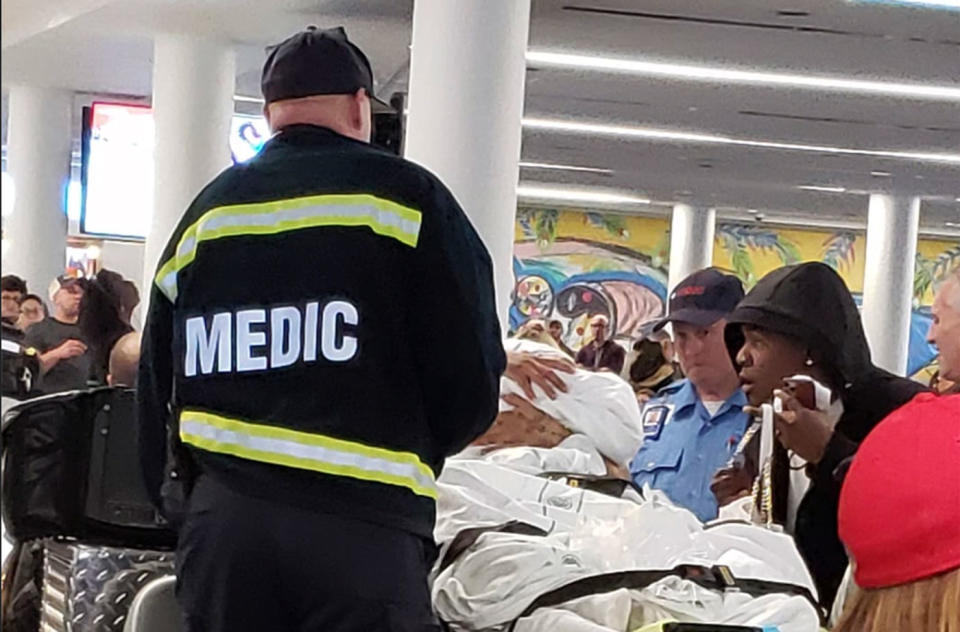 A paramedic looks on at a woman in a stretcher who's just had a baby. They're inside an airport. Bystanders also look on.