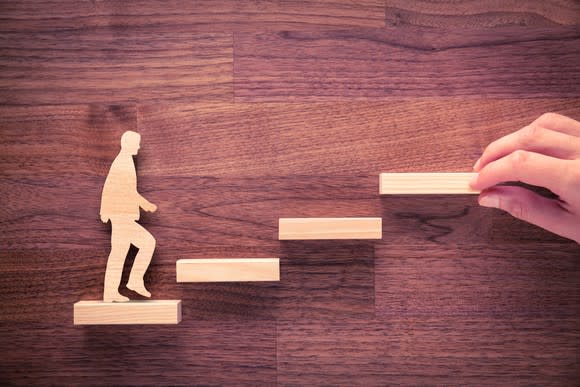 wooden cutout of a man stepping up wood block steps. A human hand assemples the pieces from the right side.