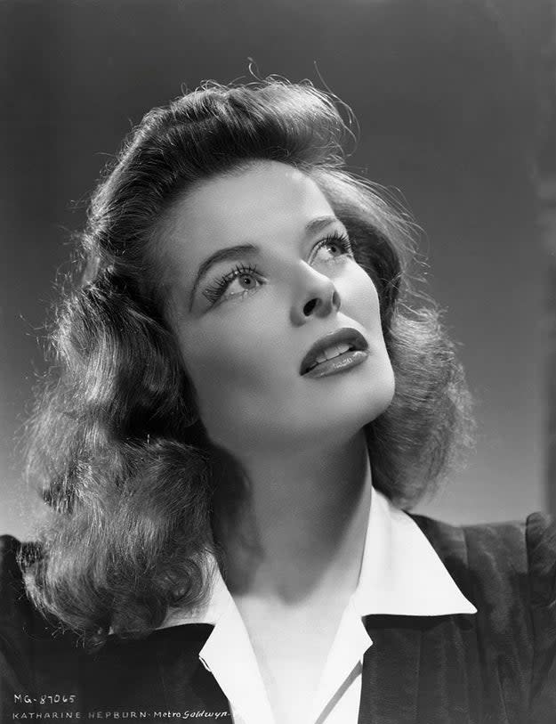 Katharine Hepburn looks upwards with a thoughtful expression. She is wearing a collared blouse and a black jacket