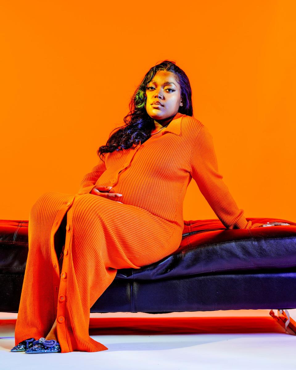 Anifa Mvuemba poses in an orange knit dress and purple stiletto shoes against a neon orange backdrop.