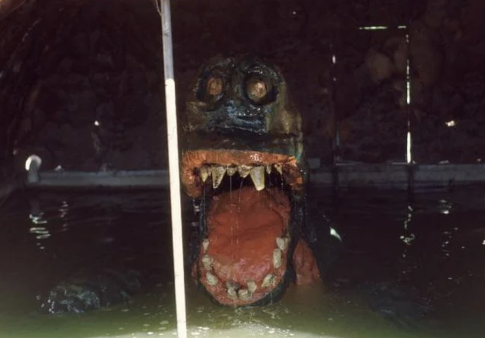 A statue of a large, open-mouthed creature with sharp teeth submerged in water, resembling a crocodile or dinosaur, found in an unusual setting