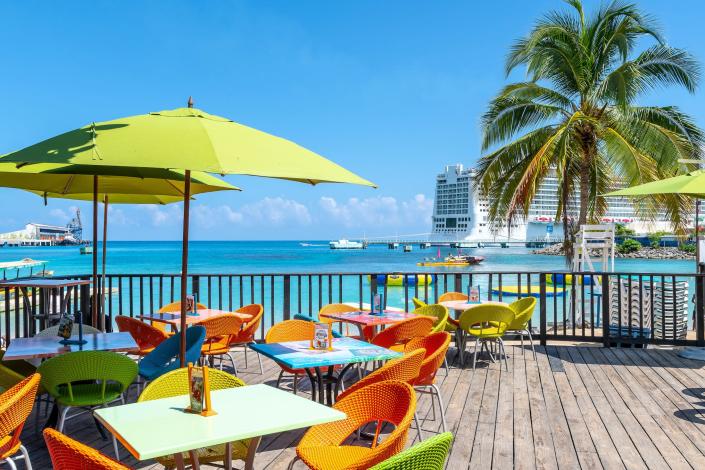 Colorful chairs and umbrellas on a wooden terrace with a view of the sea, a palm tree and a cruise ship