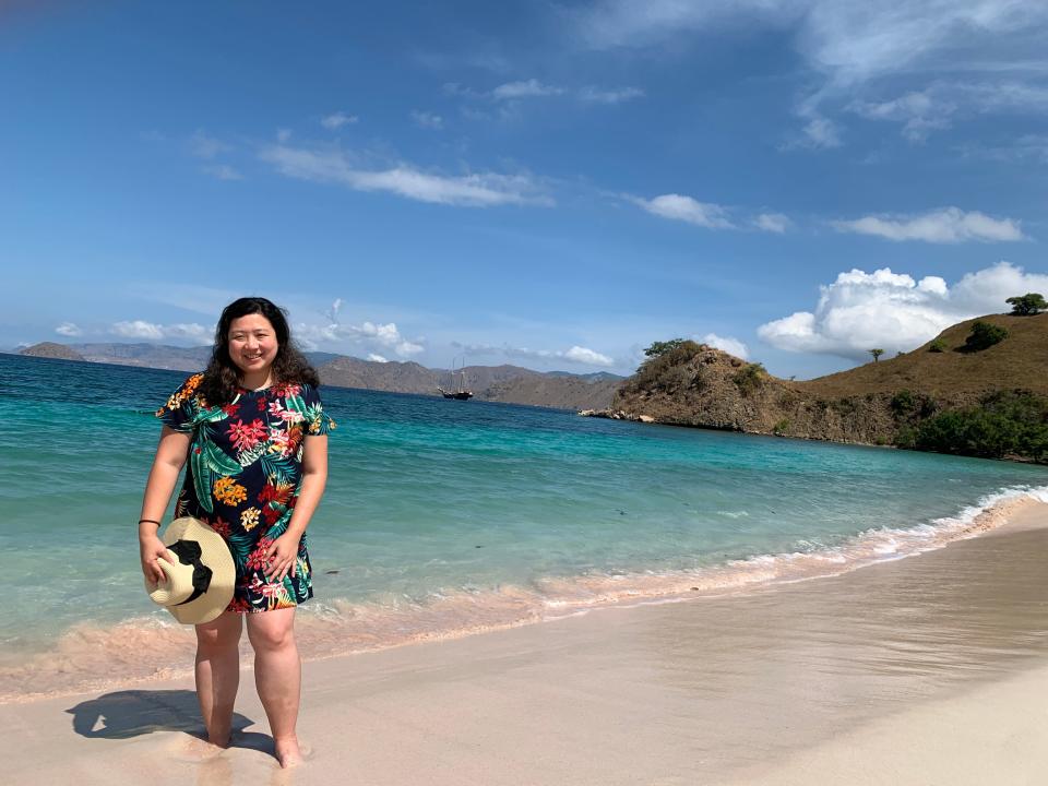 A woman posing in front of a tropical beach holding her sunhat.